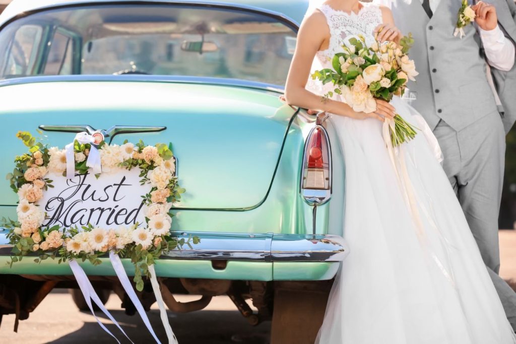 newlyweds posing by a car with a floral sign that says just married