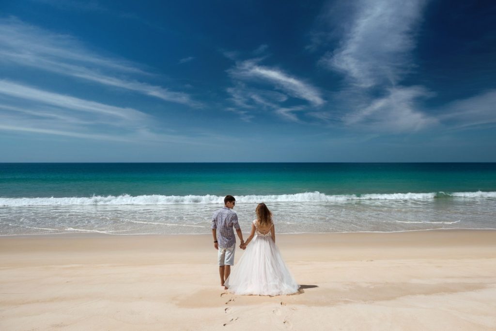 couple in wedding attire holding hands on the beach facing the ocean